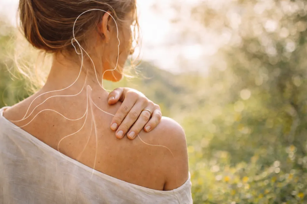 Mulher de costas ao ar livre, com luz natural e vegetação desfocada ao fundo, transmitindo bem-estar e serenidade